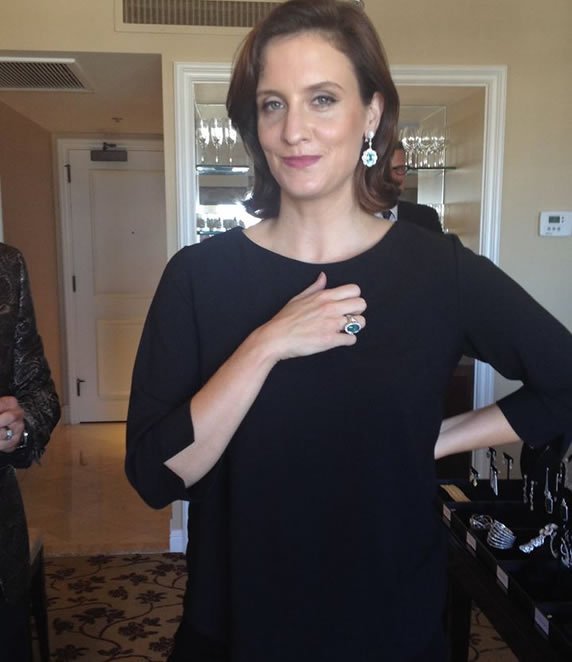 A woman with short, wavy auburn hair wearing a black dress and large earrings, standing indoors with a mirror behind her and jewelry on display to her side.