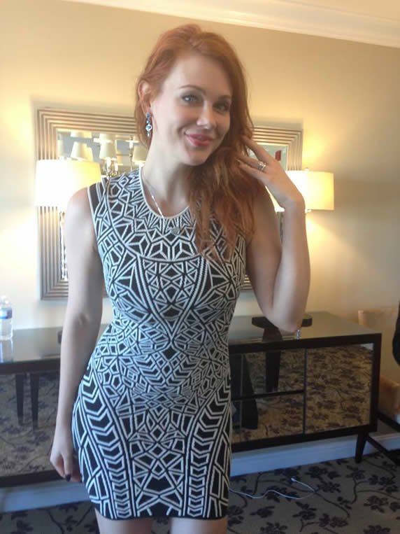 A woman with red hair and fair skin wearing a sleeveless, black-and-white geometric patterned dress inside a room with warm lighting, a mirror, lamps, and a dark table.