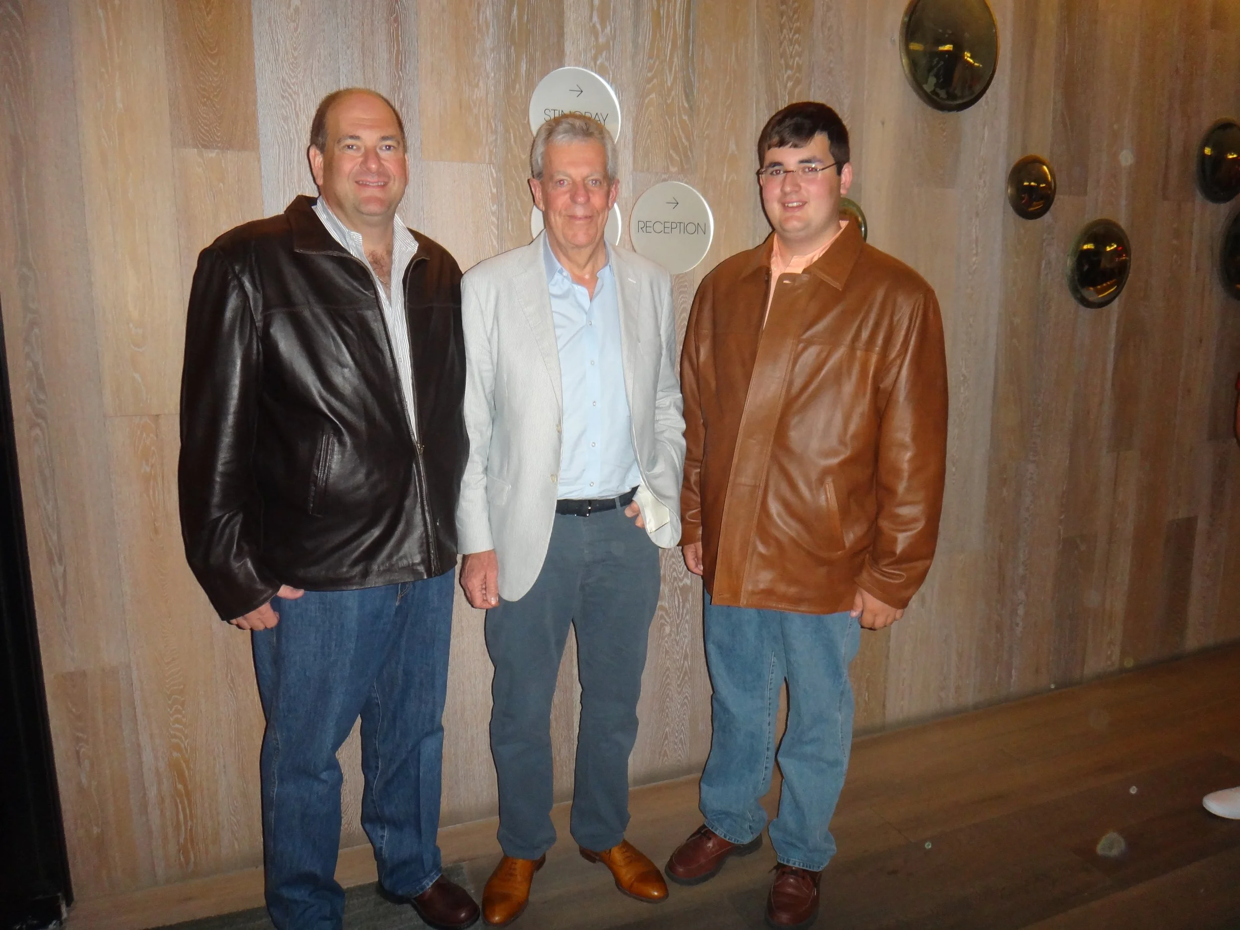 Three men standing together indoors in front of a wood-paneled wall, smiling at the camera.