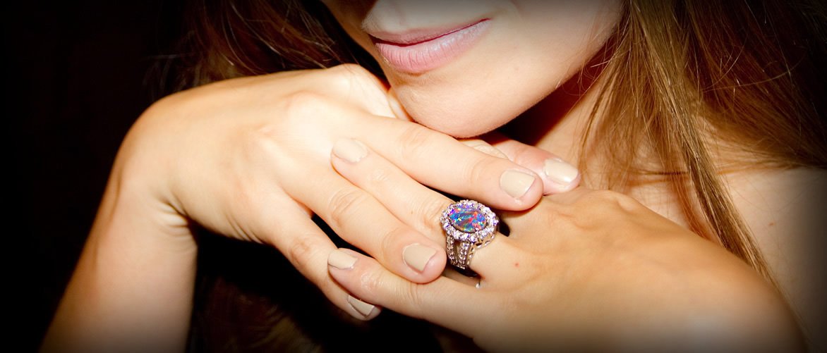 Close-up of a woman with a large, colorful gemstone ring on her finger, her hands gently touching her chin and lips, with a soft smile and reddish hair.