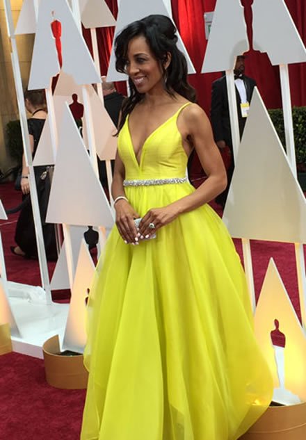 Woman in a bright yellow gown holding a small clutch, standing at an awards event with red carpet and Oscar statue cutouts in the background.