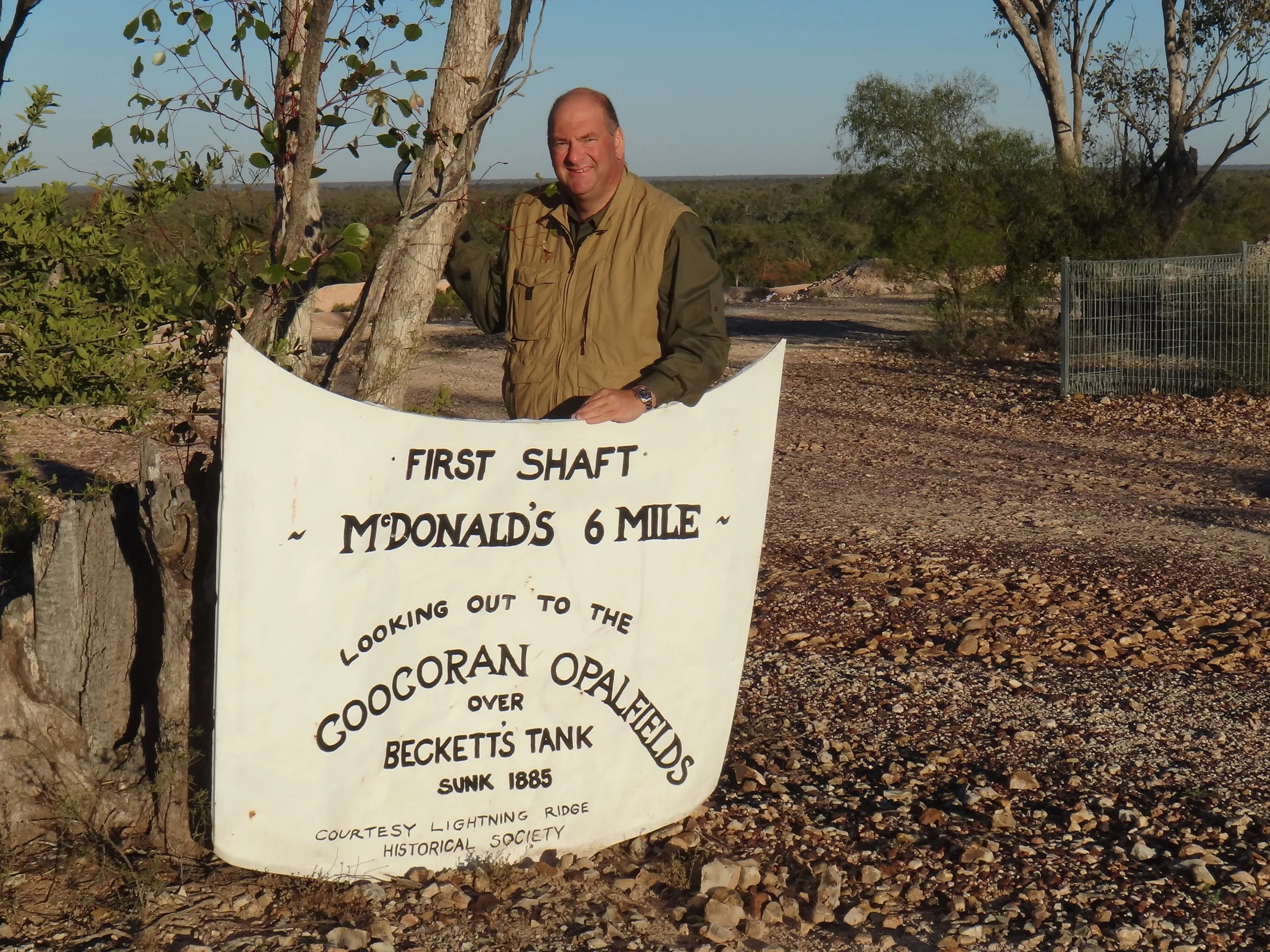 A man standing behind a white sign about McDonald's 6 miles away, with desert trees and gravel road in background.