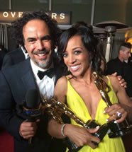 A man and woman smiling and holding wine glasses at an event, with the man wearing a tuxedo and the woman in a yellow dress.