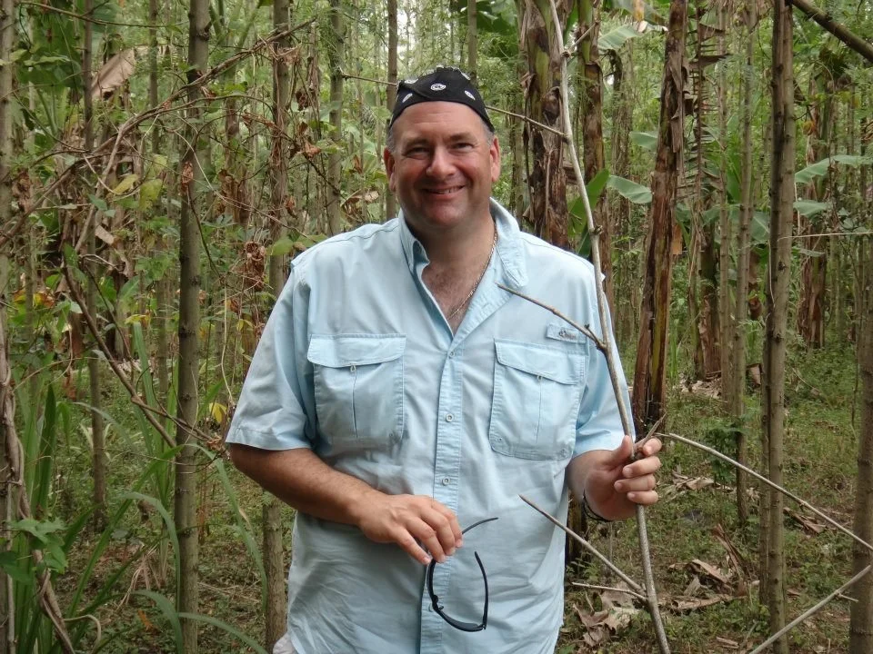 A smiling man wearing a light blue short-sleeved shirt and a black bandana stands in a dense forest, holding a small branch in his right hand and sunglasses in his left hand.