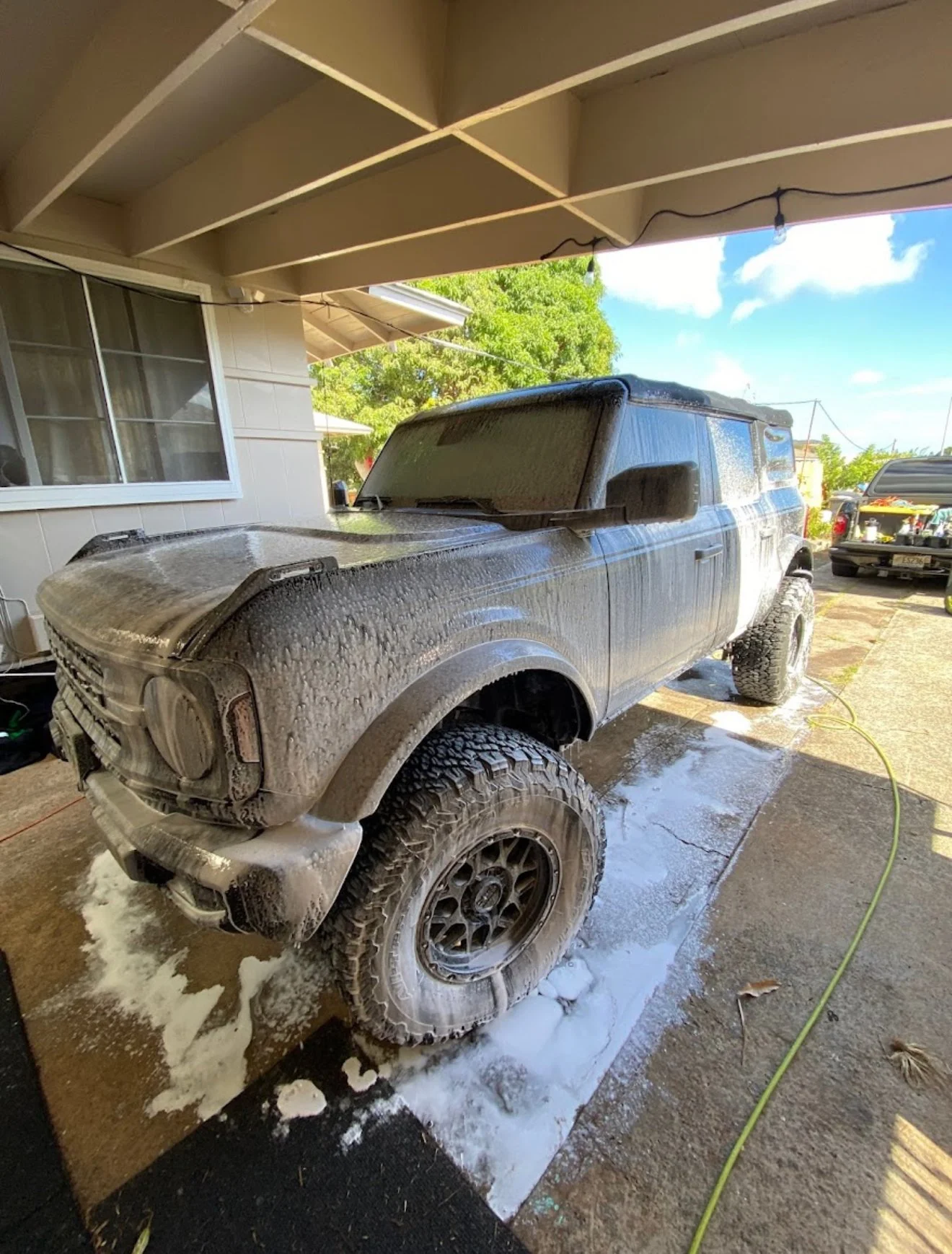 A black off-road vehicle covered in soap suds during a wash, parked on a concrete driveway under a beige awning, with a green tree and blue sky in the background.