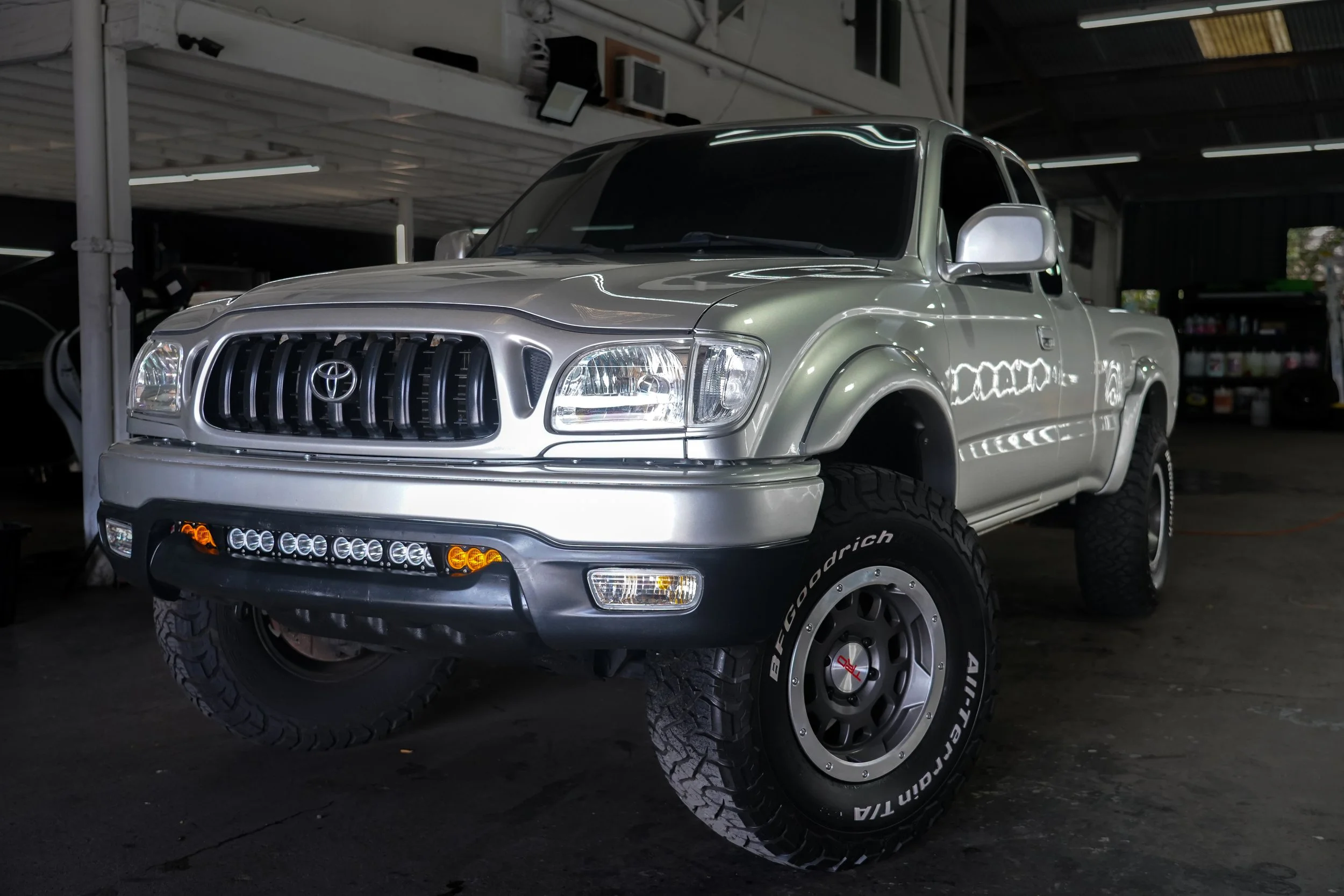 Silver Toyota pickup truck parked inside a garage with off-road tires and custom front lights.