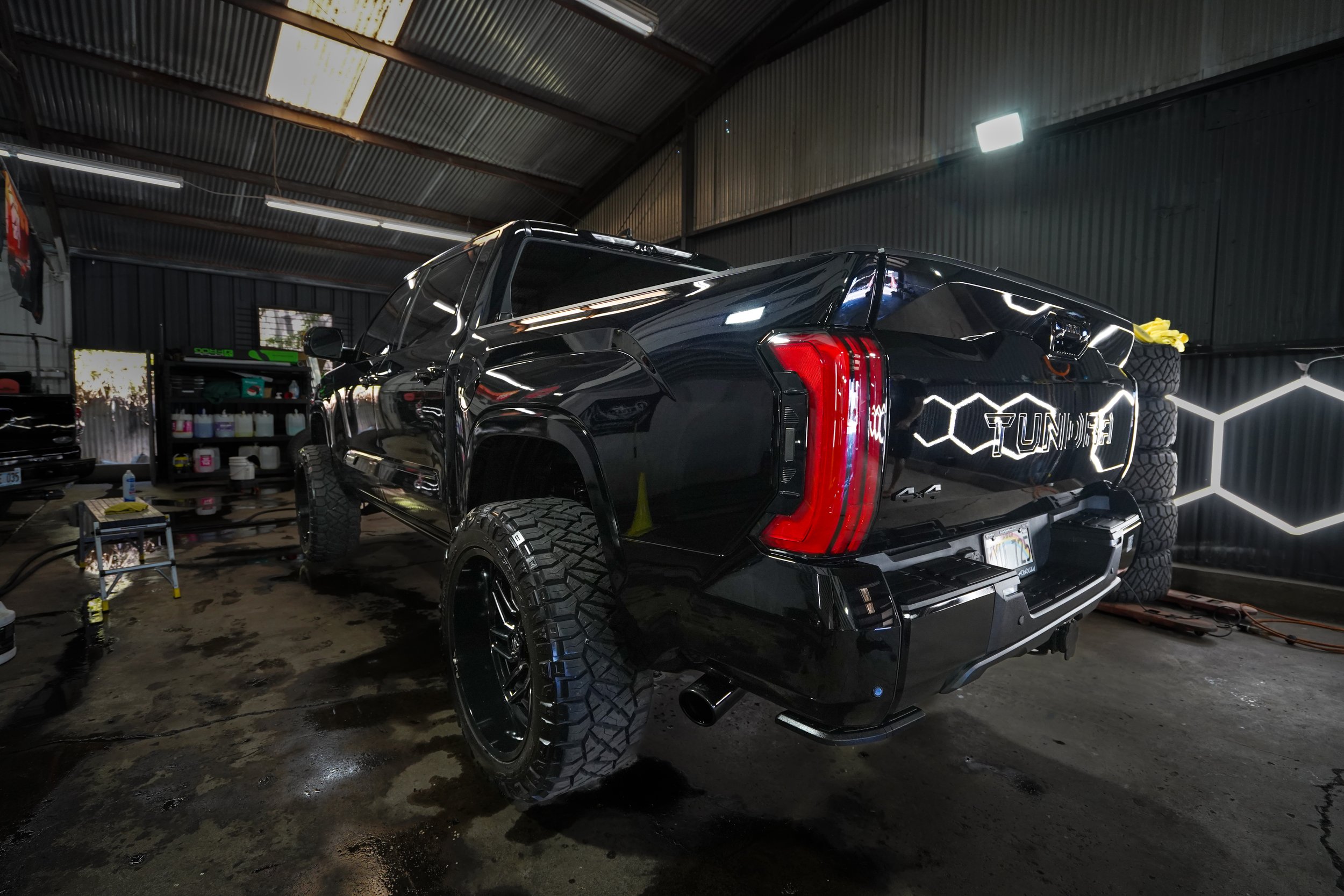 Black pickup truck inside a garage, with the word 'TUNED' on the tailgate, surrounded by automotive tools, supplies, and stacked tires.