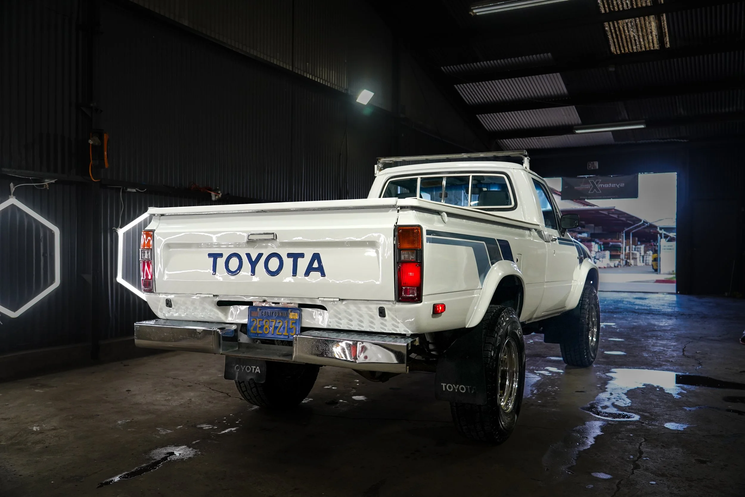 White vintage Toyota pickup truck inside a garage, with the brand name 'TOYOTA' visible on the tailgate, and a California license plate. The garage has black walls with hexagonal light fixtures and an open garage door in the background.