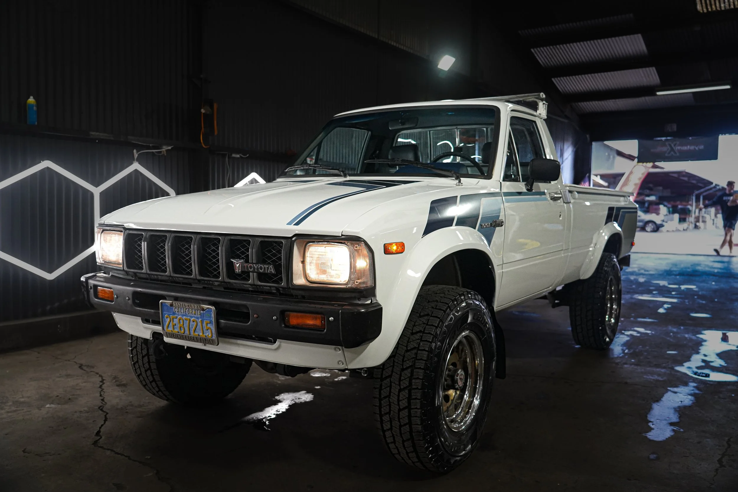 White vintage Toyota pickup truck inside a dark garage with a dark wall and neon hexagon designs, wet floor, and a person in the background.