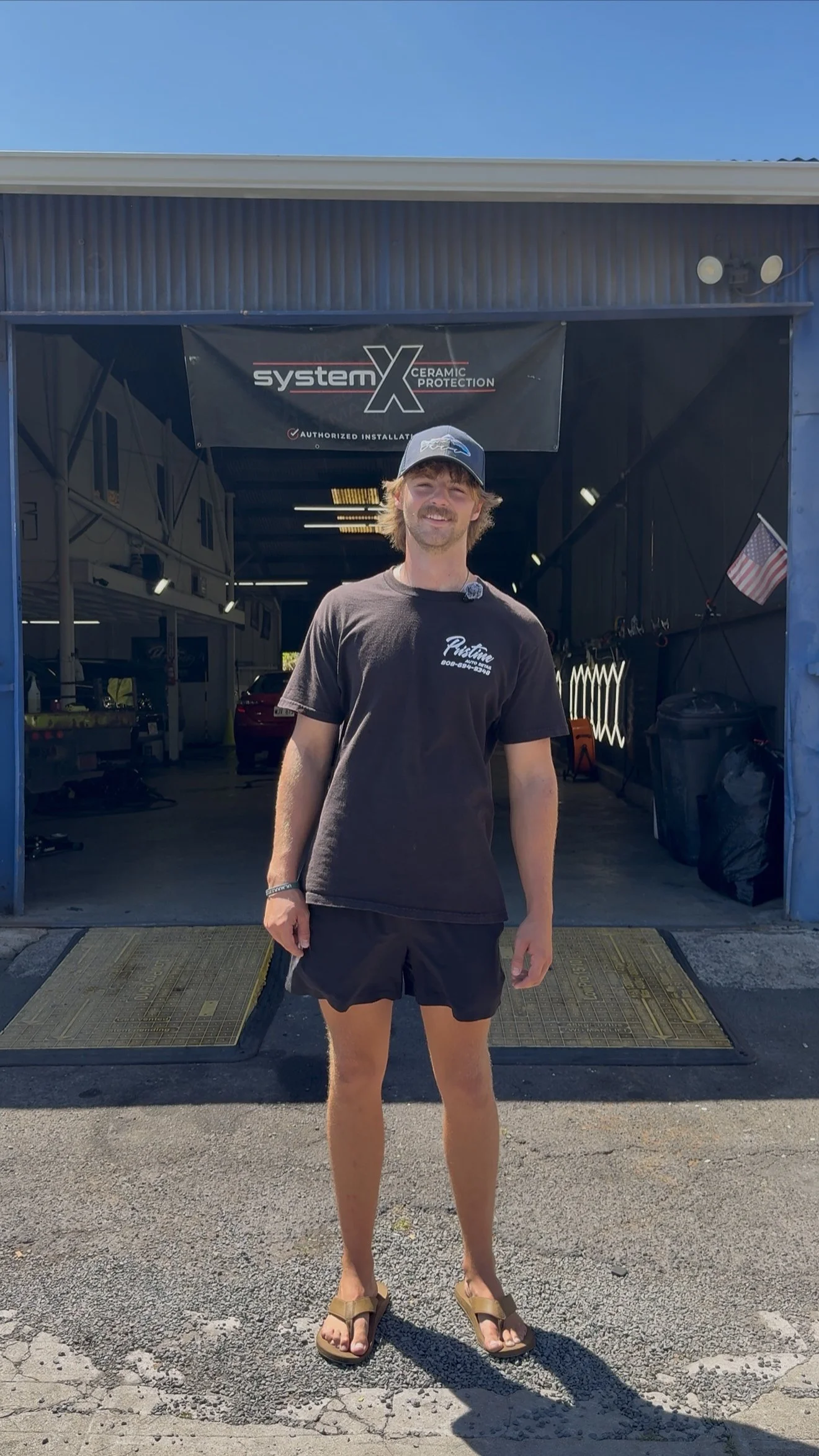 A young man with shoulder-length blond hair wearing a baseball cap, t-shirt, shorts, and flip-flops standing outside a garage with an open door. Inside the garage are vehicles, tools, and a sign reading 'systemX Ceramic Protection'.