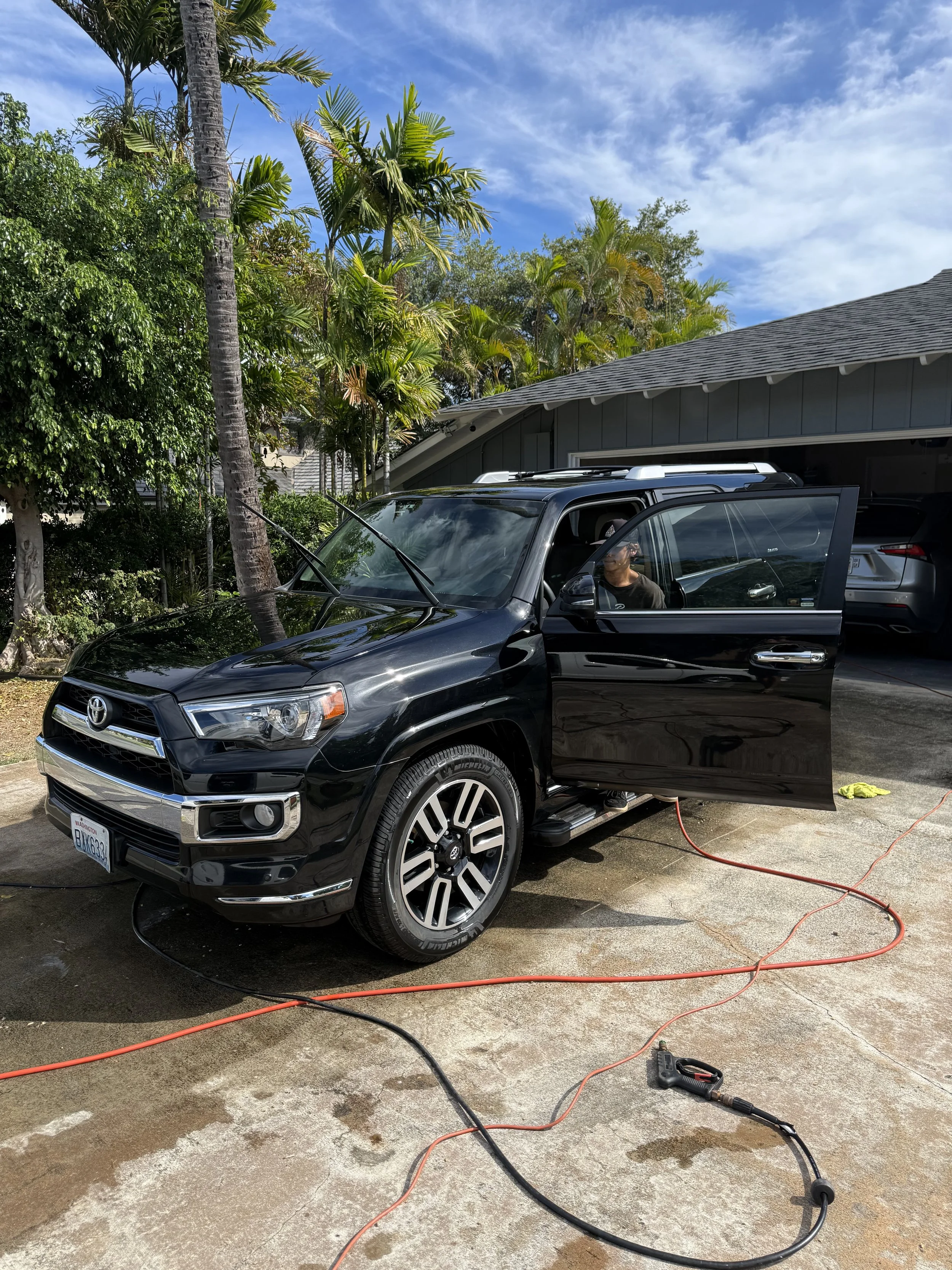 A black Toyota SUV parked outside a house, with the driver's side door open. There are electrical cords connected to the vehicle, possibly for charging or maintenance. The scene is sunny with a clear blue sky and lush palm trees in the background.