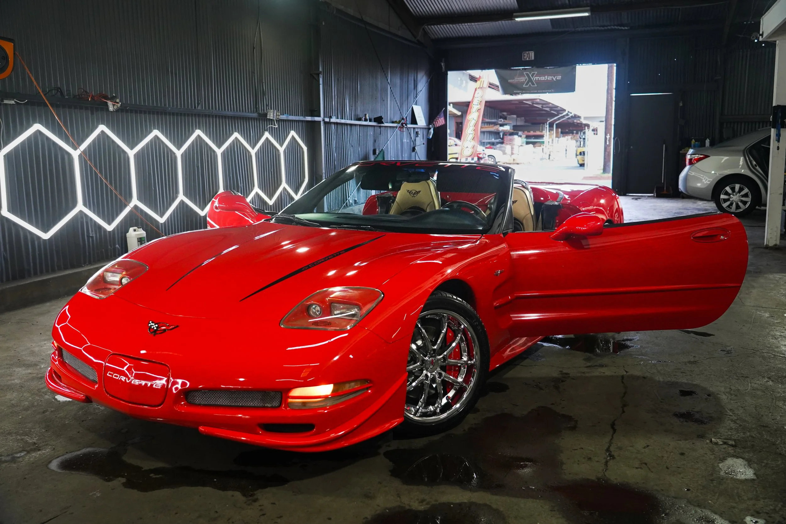 Red Chevrolet Corvette convertible inside a garage with one door open, showing bright sunlight outside, with a white car parked to the right.