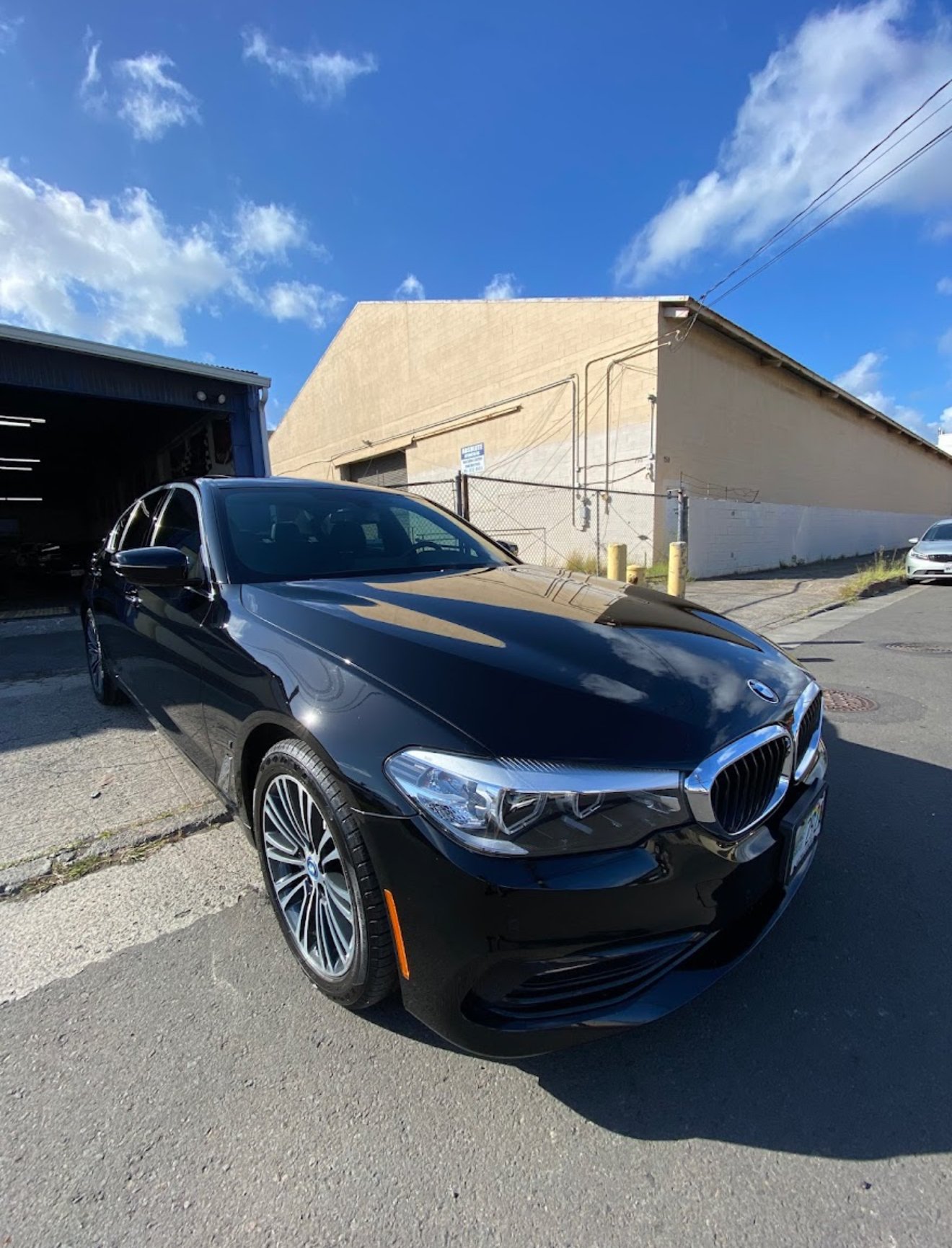 Black BMW sedan parked outdoors in front of a beige building with a bright blue sky and scattered clouds overhead.