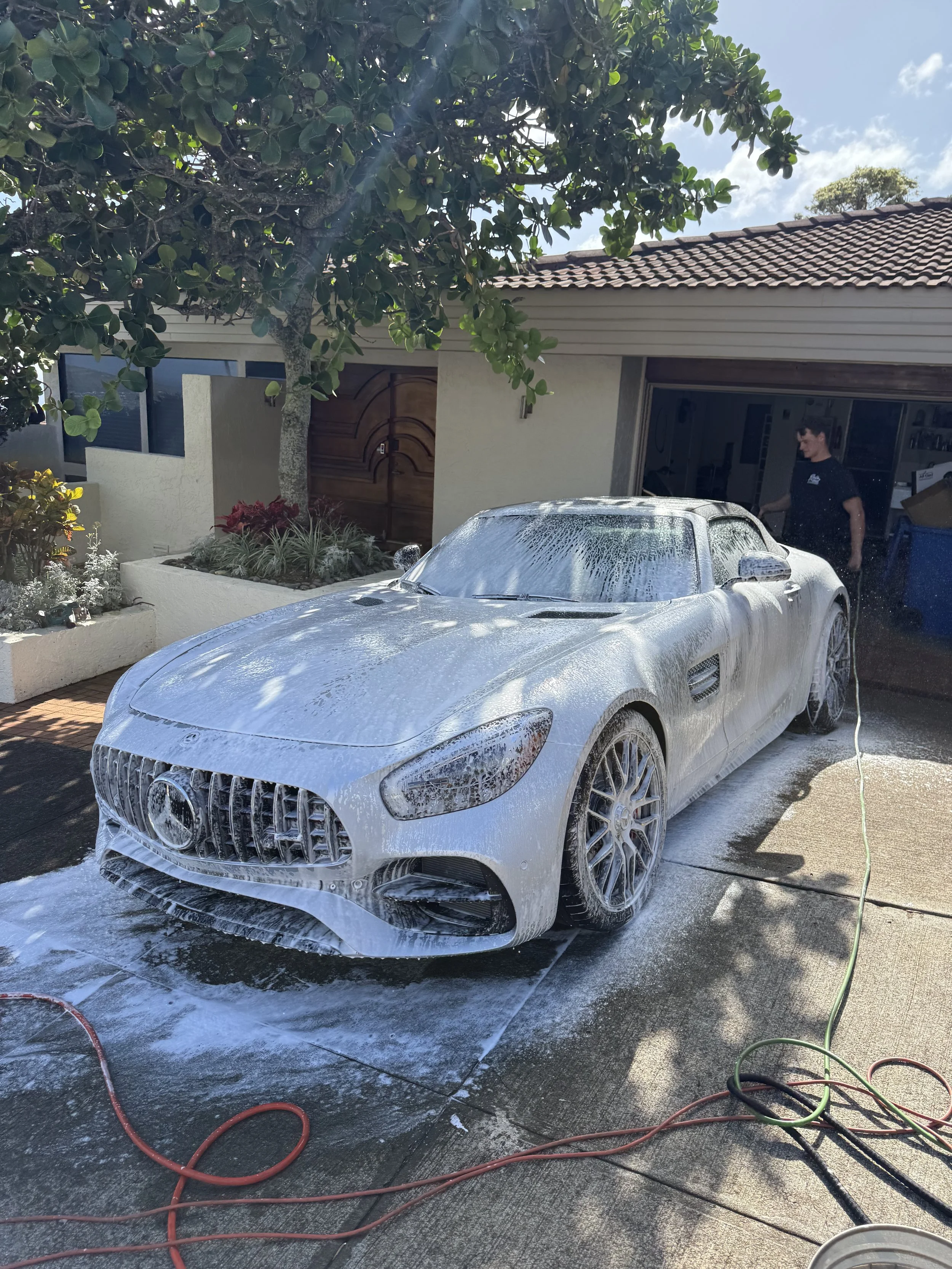 A white Mercedes-Benz sports car being washed with soap and foam in a driveway under a tree, with a person in a black shirt standing nearby.