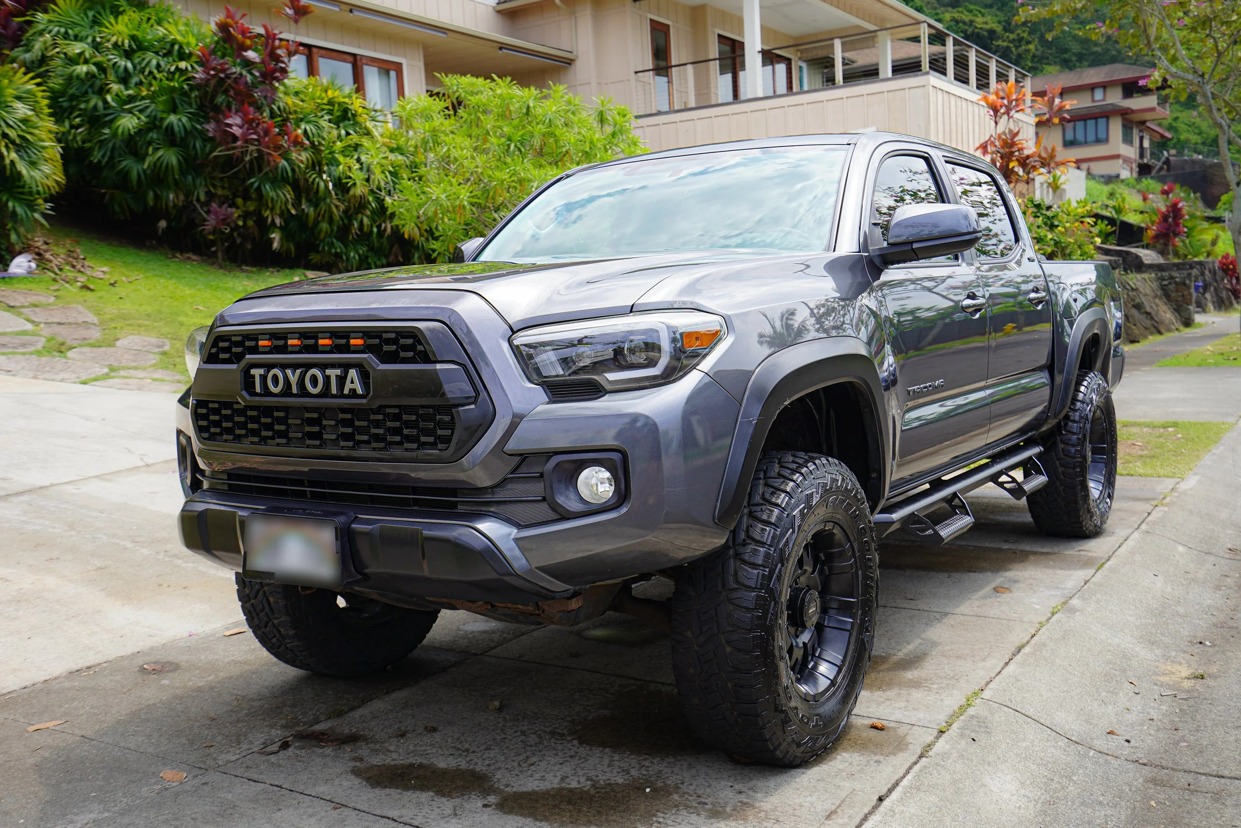 Gray Toyota Tacoma pickup truck parked on a driveway in front of a house with lush green and colorful landscaping.
