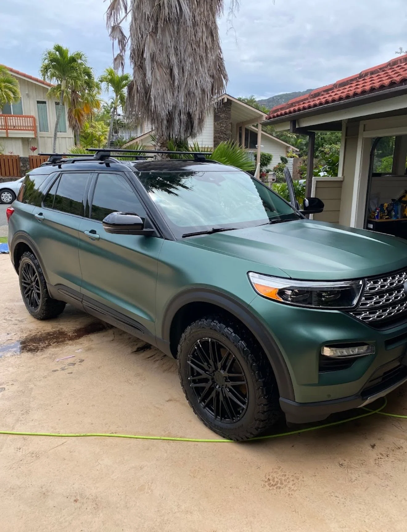 Green SUV parked on driveway in front of a house with palm trees and a large palm tree nearby.