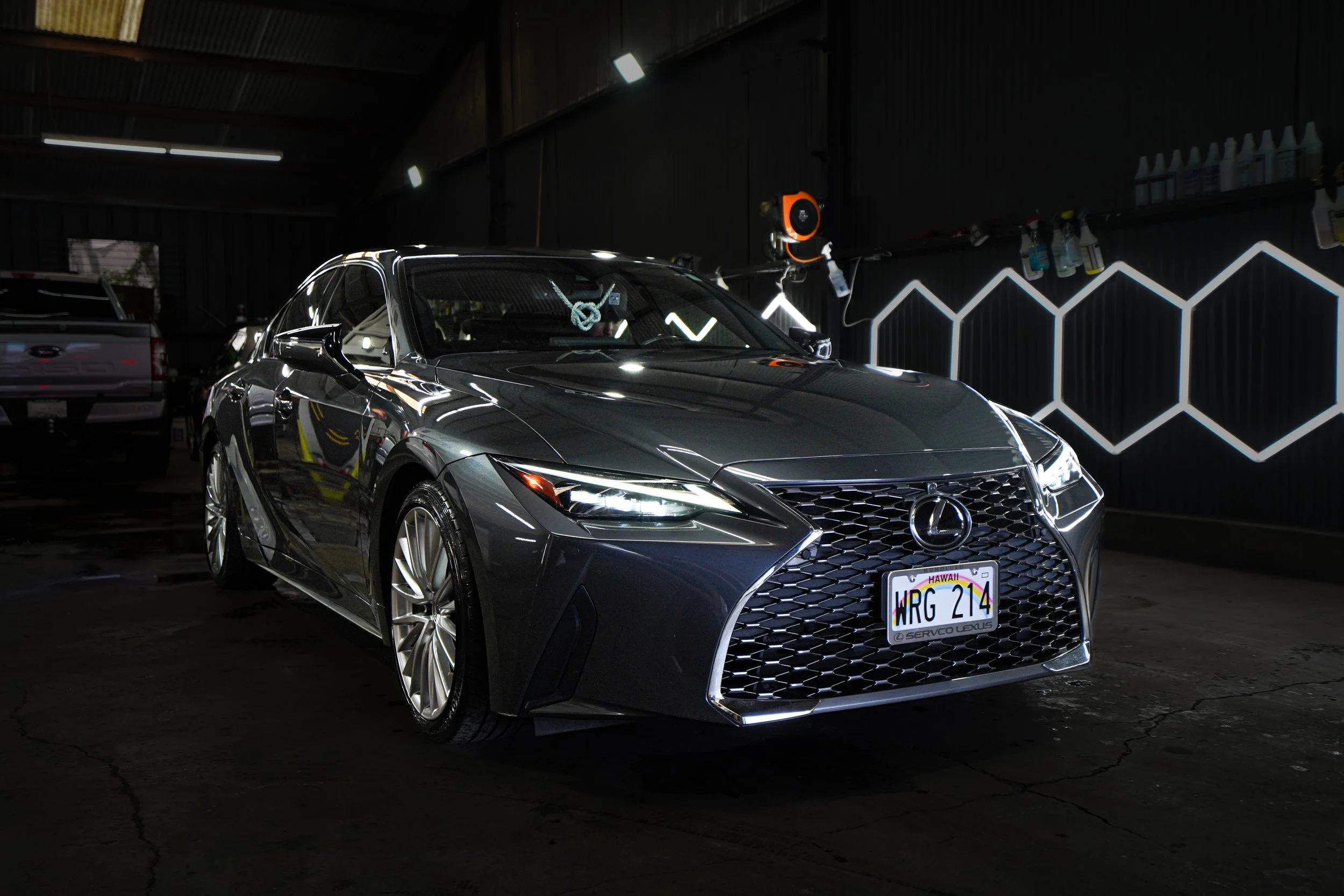 A dark gray Lexus sedan in a dimly lit car wash or detailing shop with hexagonal LED light designs on the wall in the background.