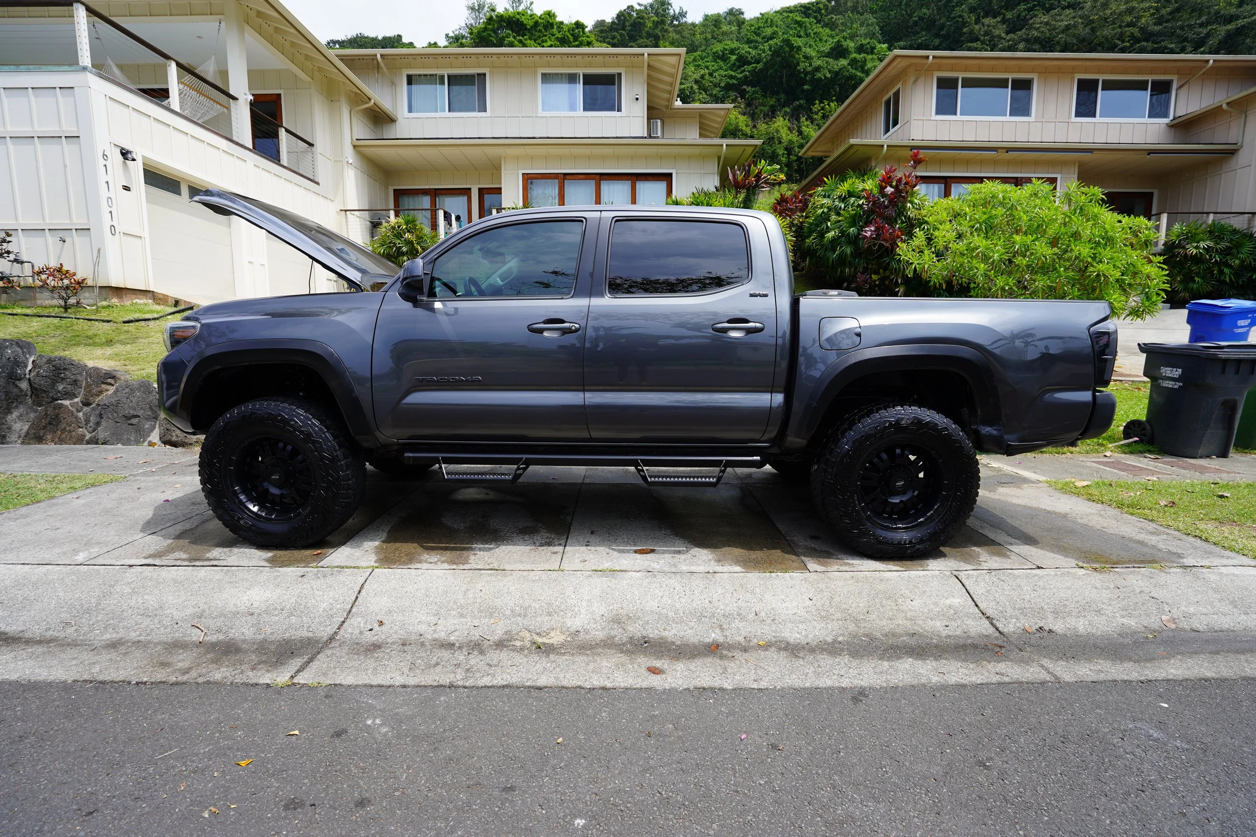 A dark gray Toyota Tacoma pickup truck with its hood open, parked in a residential driveway. The truck has large off-road tires and black rims. In the background, there are modern two-story yellow houses with balconies and greenery.
