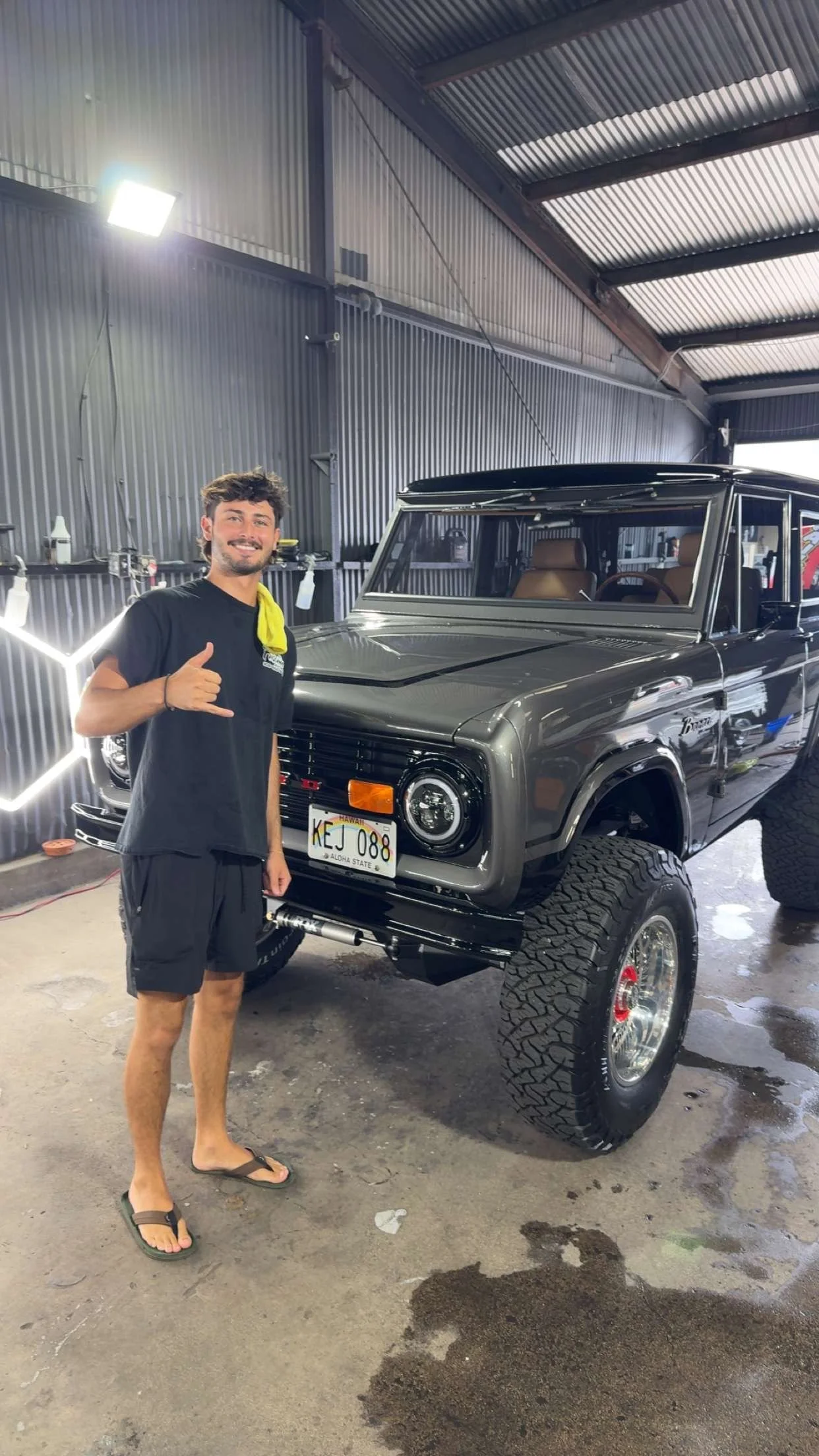 A young man smiling and making a shaka hand gesture next to a black vintage off-road vehicle in a garage or workshop. The vehicle has large tires, a listings plate from Hawaii, and interior visible through windows.