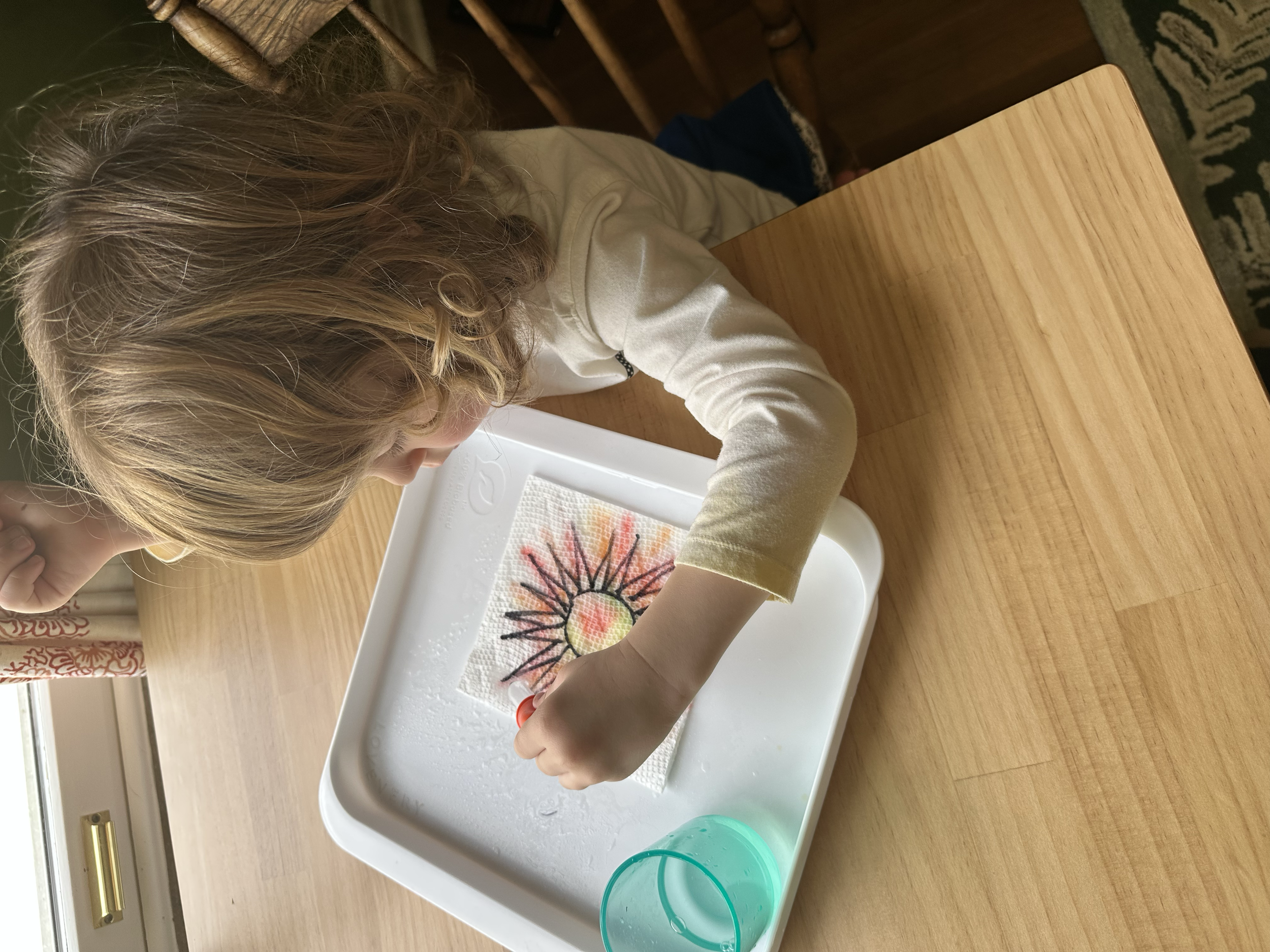 Child creating a sand art picture of a sun on a paper towel, with water and a tray, sitting at a wooden table.