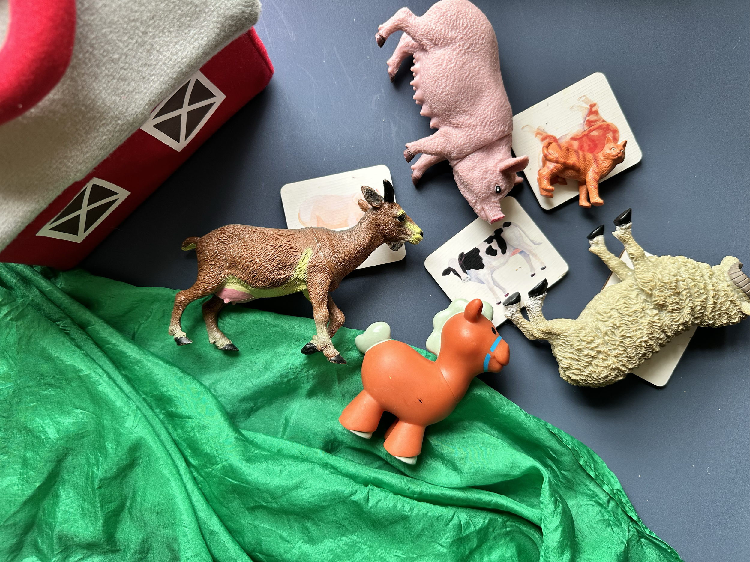 Collection of small animal figurines including a pink pig, a brown donkey, a cream-colored sheep, an orange horse, and two animal cards, with a green fabric and a red and white barn-themed box nearby.