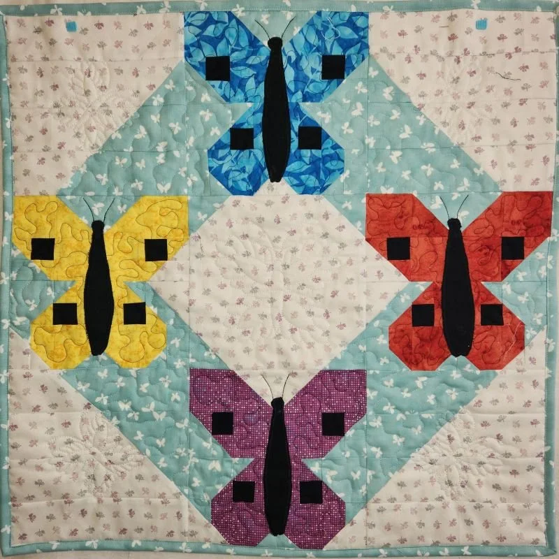 Colorful quilted wall hanging featuring four butterfly designs in blue, yellow, red, and purple arranged in a geometric pattern on a light patterned background