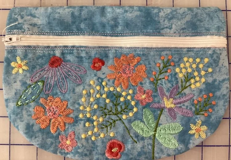 Hand-embroidered floral zipper pouch created in embroidery class featuring detailed stitched flowers on blue fabric