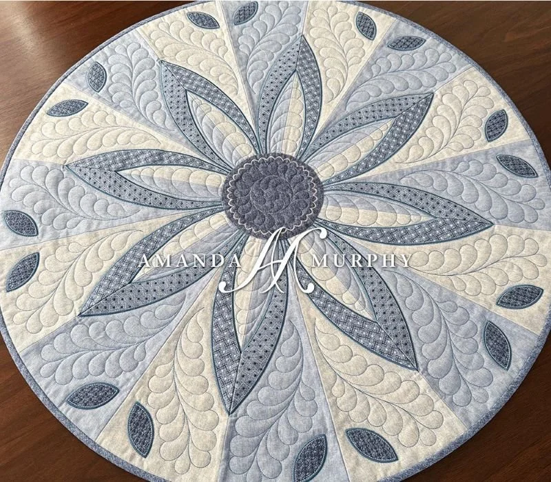 Circular quilted table topper featuring a large blue and gray floral design with pointed petals, intricate stitching, and a patterned cream background.