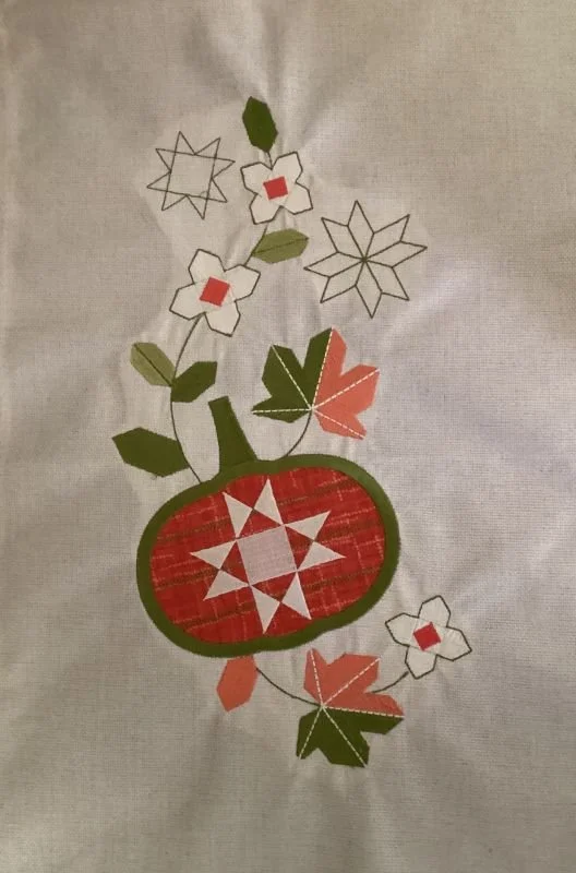 Embroidered fall table runner design featuring a red pumpkin surrounded by green leaves, delicate floral accents, and autumn-inspired appliqué on cream fabric.