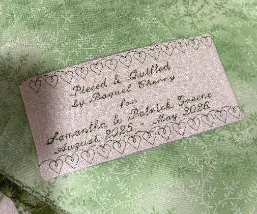 Personalized rectangular fabric quilt label with embroidered lettering and a decorative stitched border, displayed on green quilted fabric.