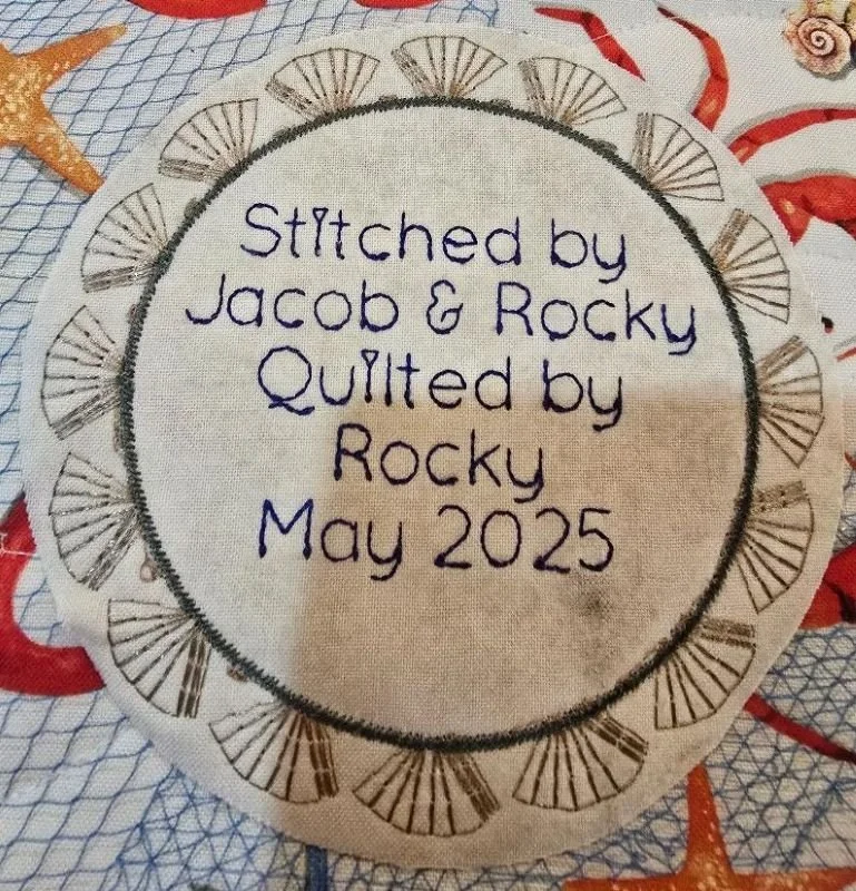 Circular embroidered quilt label featuring blue handwritten-style lettering reading "Stitched by Jacob & Rocky, Quilted by Rocky, May 2025," surrounded by a decorative patterned fabric border.