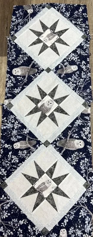 Quilted winter table runner featuring three white diamond-shaped blocks with gray and navy blue patchwork stars, surrounded by dark blue floral fabric