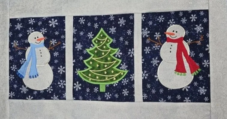 Winter embroidered appliqué project featuring three navy blue fabric panels with two white snowmen wearing colorful scarves and a decorated green Christmas tree.
