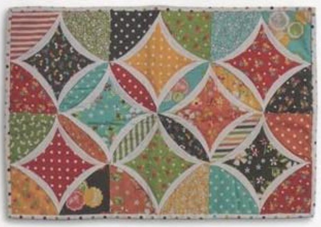 Colorful quilted Cathedral Window table mat featuring curved white fabric frames surrounding geometric patchwork squares in red, green, teal, yellow, and floral prints.