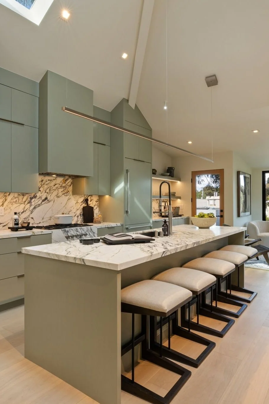 Modern kitchen with marble island, bar stools, and green cabinets. High ceiling with recessed lighting.