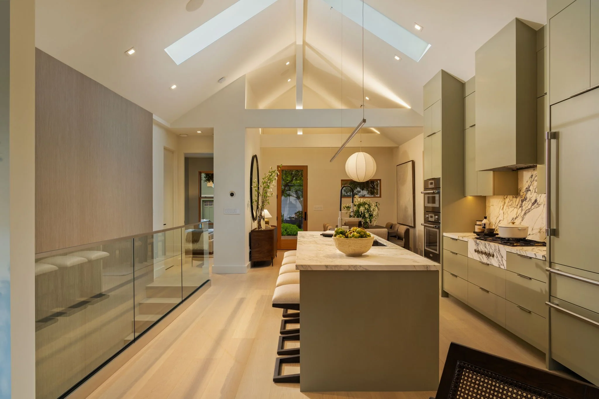 Modern kitchen with high ceilings, skylights, marble countertops, green cabinetry, stainless steel appliances, and a central island with bar stools.