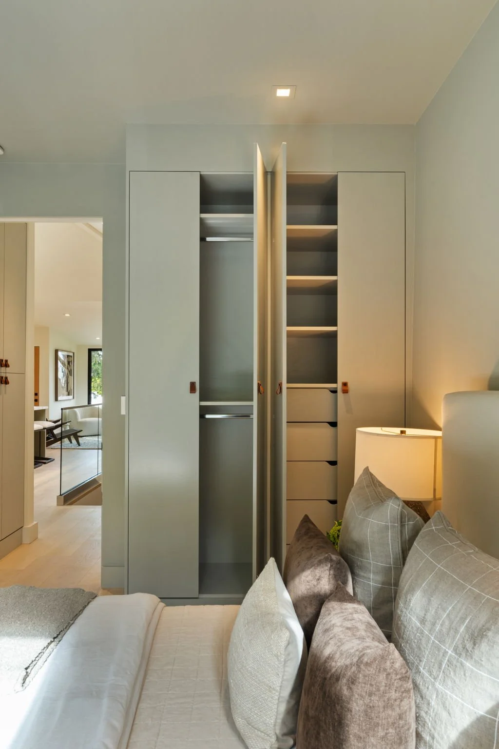 Modern bedroom with an open closet showing shelves and drawers, a bed with pillows, a bedside lamp, and a view into a living area.