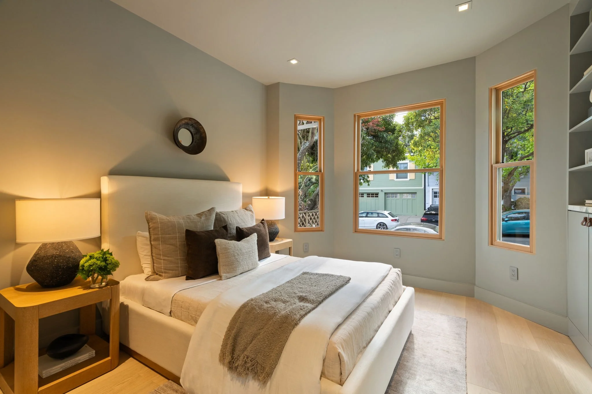 Modern bedroom interior with a neatly made bed, light wood flooring, and large windows overlooking a street with trees and parked cars. The room features a wooden side table with a lamp and decorative items, a wall mirror, and built-in shelving.