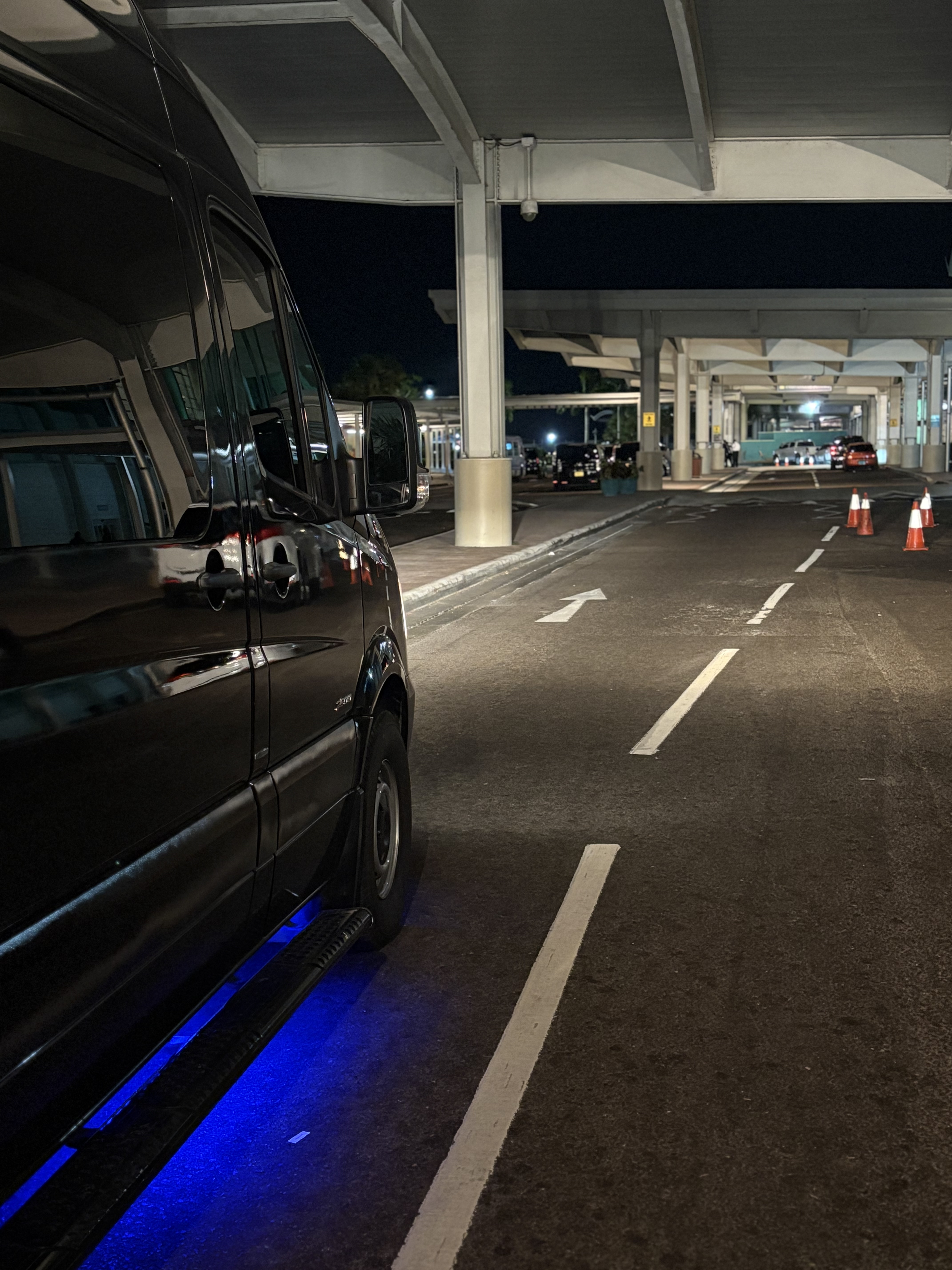 Nighttime airport drop-off area with a parked black van and blue underglow lights, reflective pavement markings and orange cones, under a roofed structure.