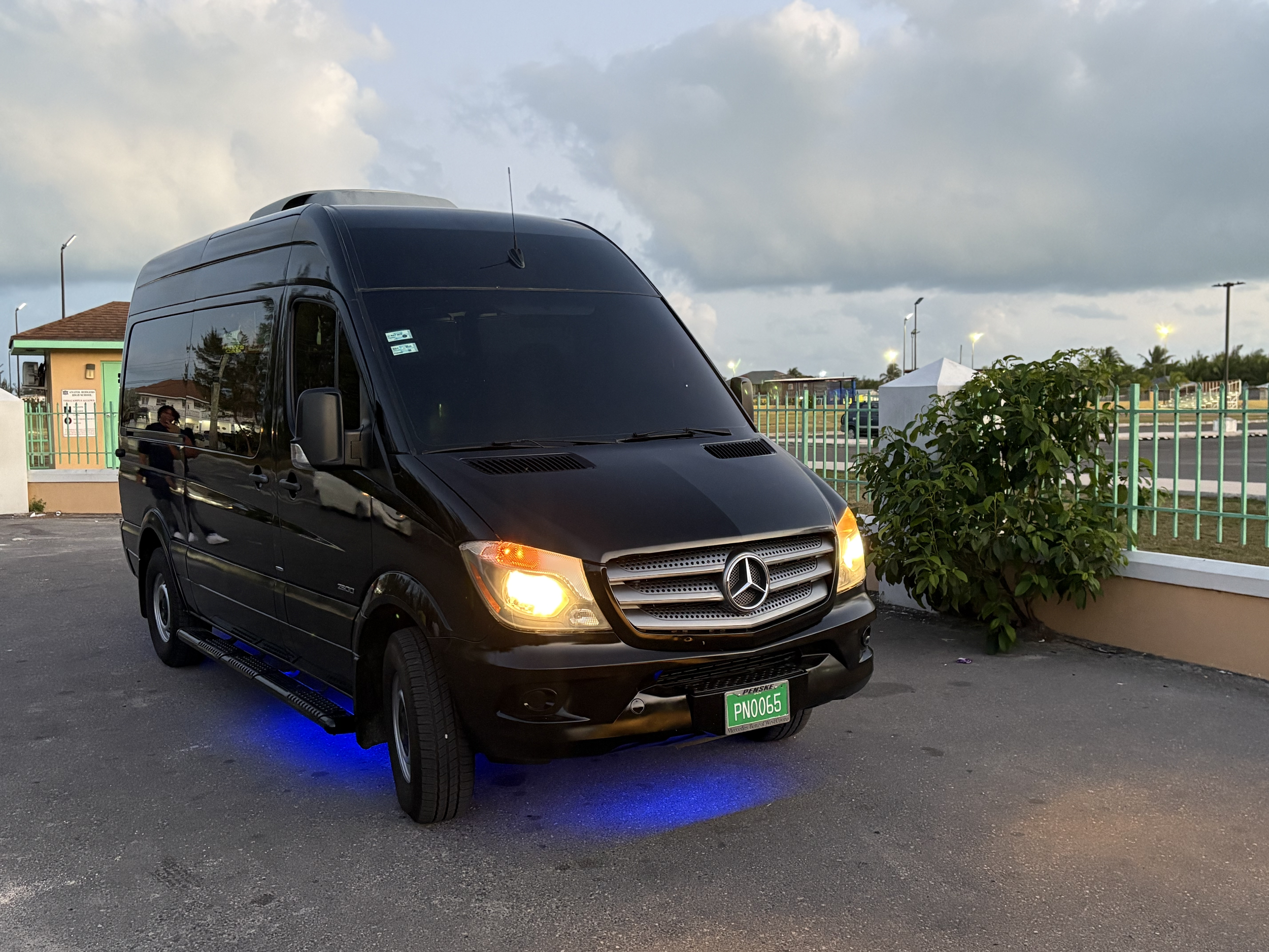 Black Mercedes-Benz van with headlights on, parked on a street, illuminated by blue underglow lights, near a building and greenery.