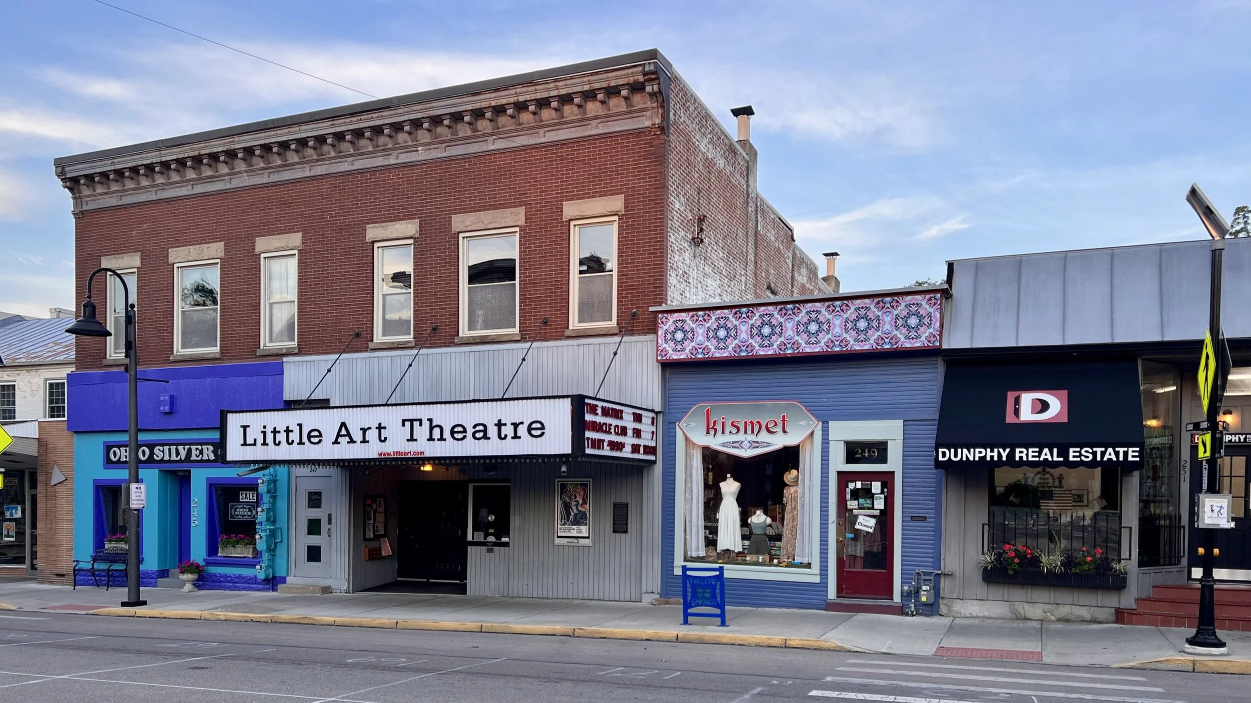 A row of downtown storefronts in Yellow Springs, Ohio, including Little Art Theatre, Kismet clothing store, and Dunphy Real Estate with a cloudy sky overhead.