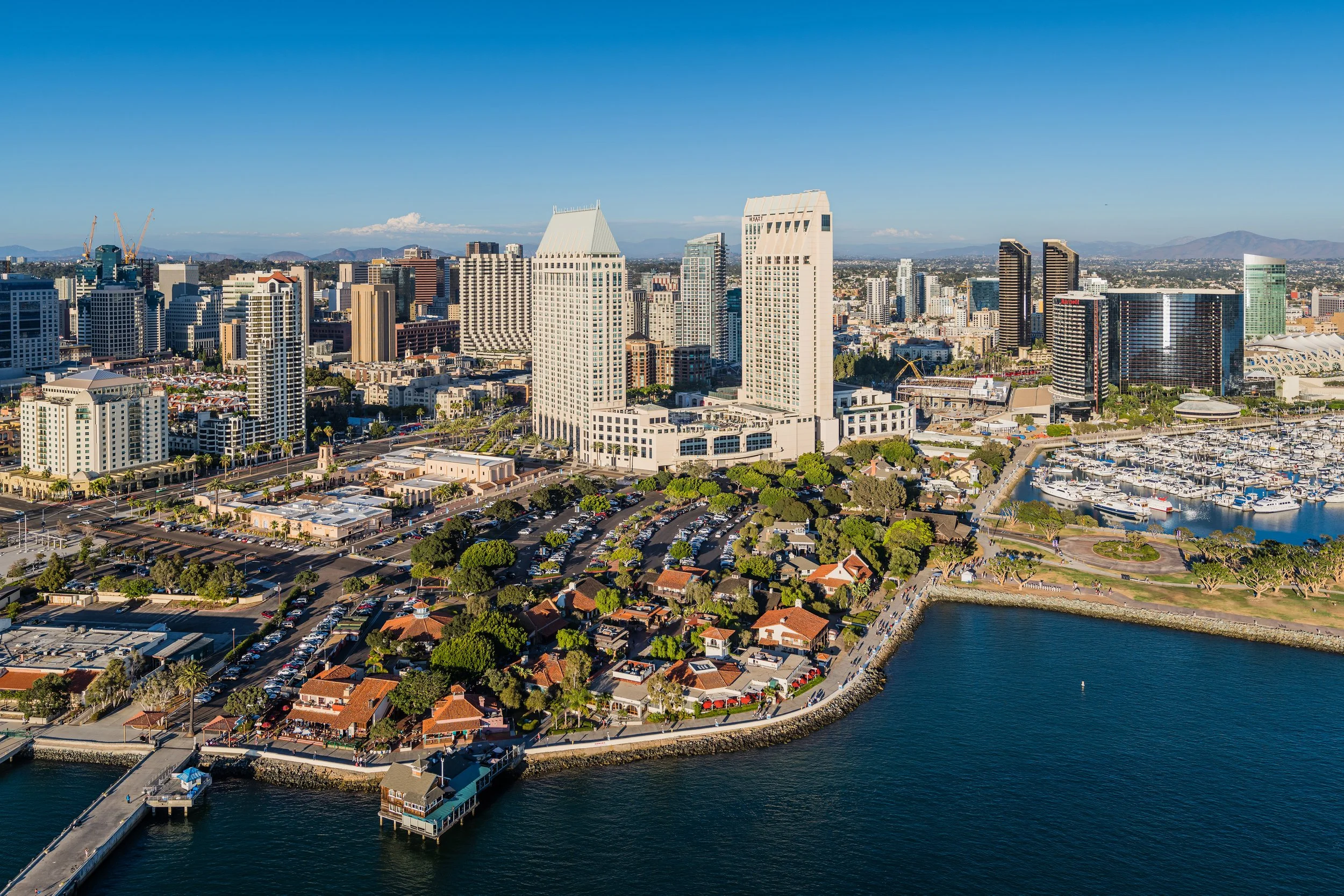 Aerial view of a city skyline with tall buildings, marina filled with boats, and a waterfront park with trees and pathways, under a clear blue sky.