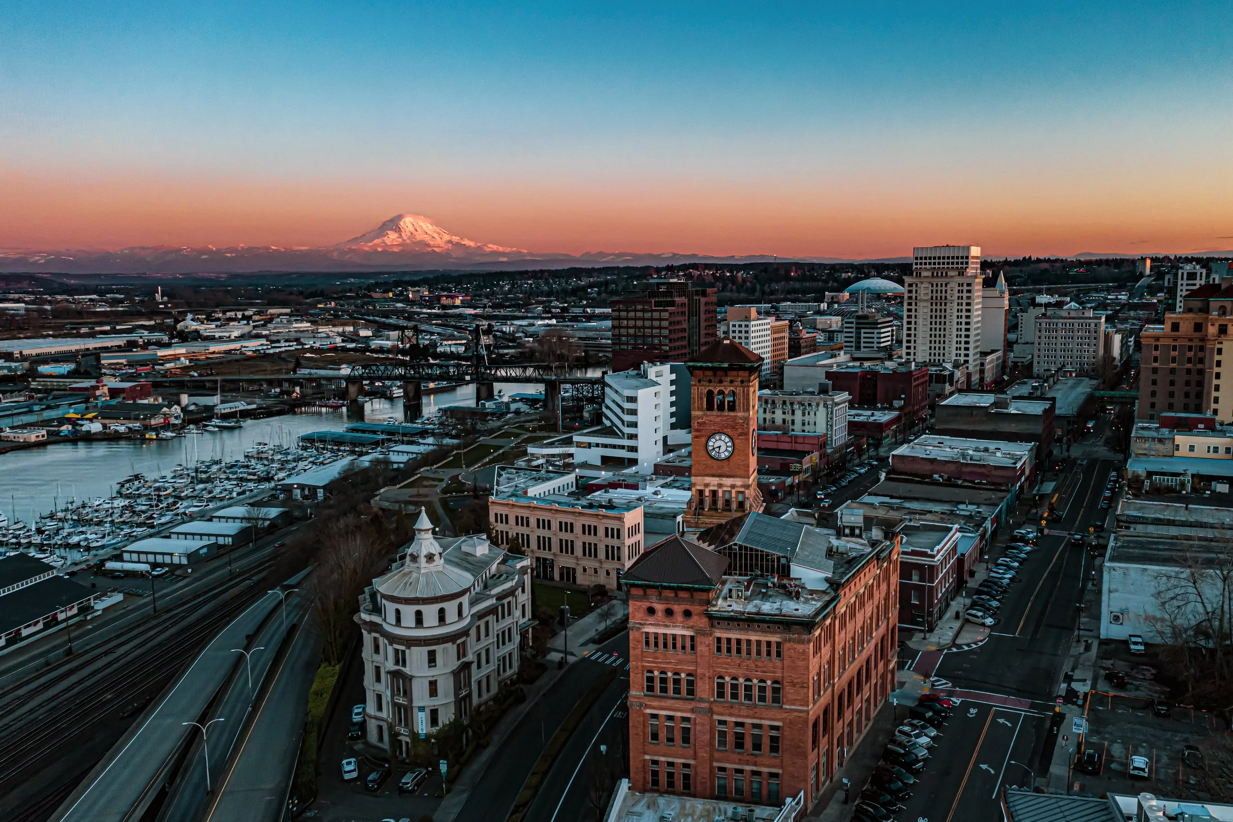 Aerial view of a cityscape at sunset with Mount Rainier in the background, featuring historic buildings, a clock tower, and a marina with boats.
