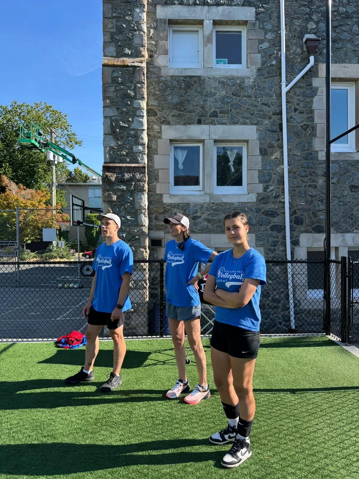 Three young volleyball players standing on a sports field with a stone building in the background, wearing blue shirts and athletic shoes.
