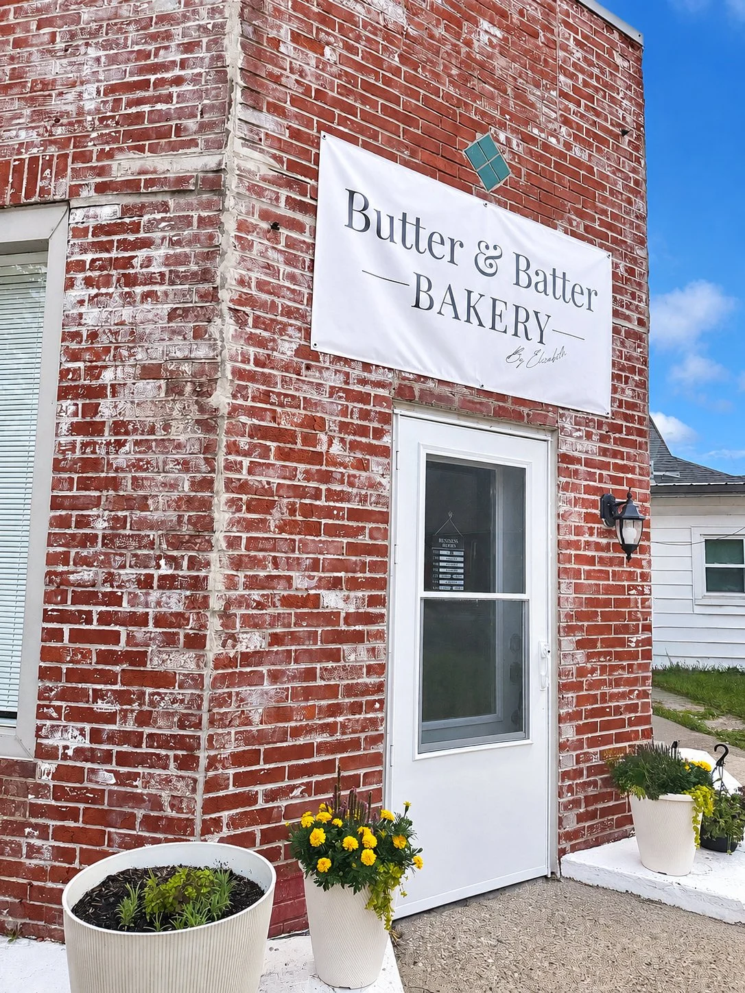 Picture of a brick building with yellow potted flowers outside. Clear blue skies in the background. Picture focuses on front door of Butter & Batter Bakery