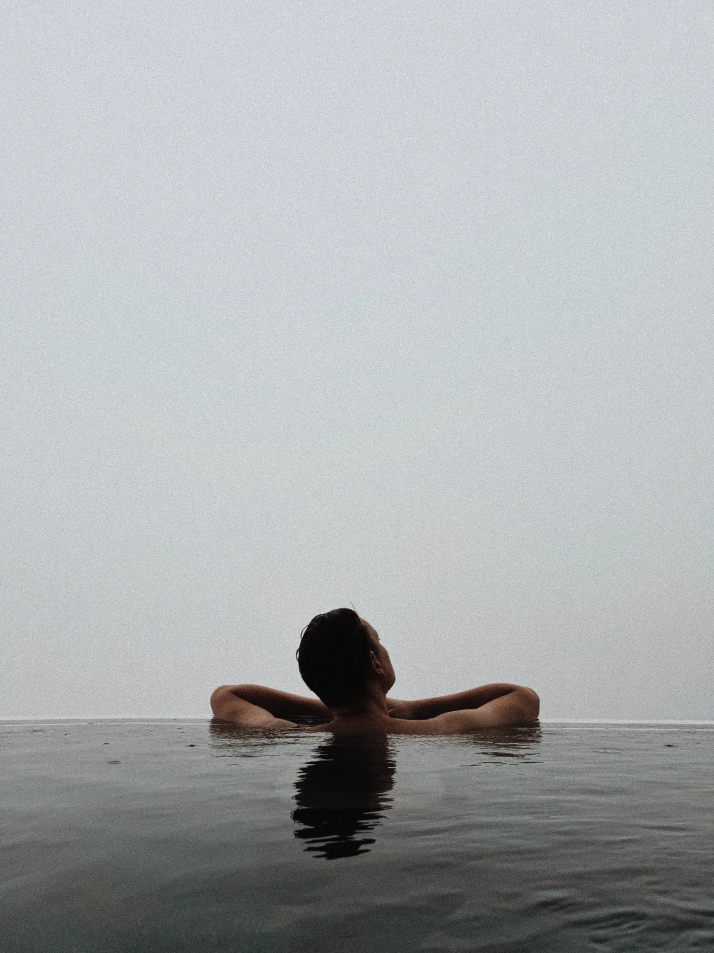 person im unendlichpool bei regen und nebel am feuerberg, wasser und himmel gehen optisch ineinander über