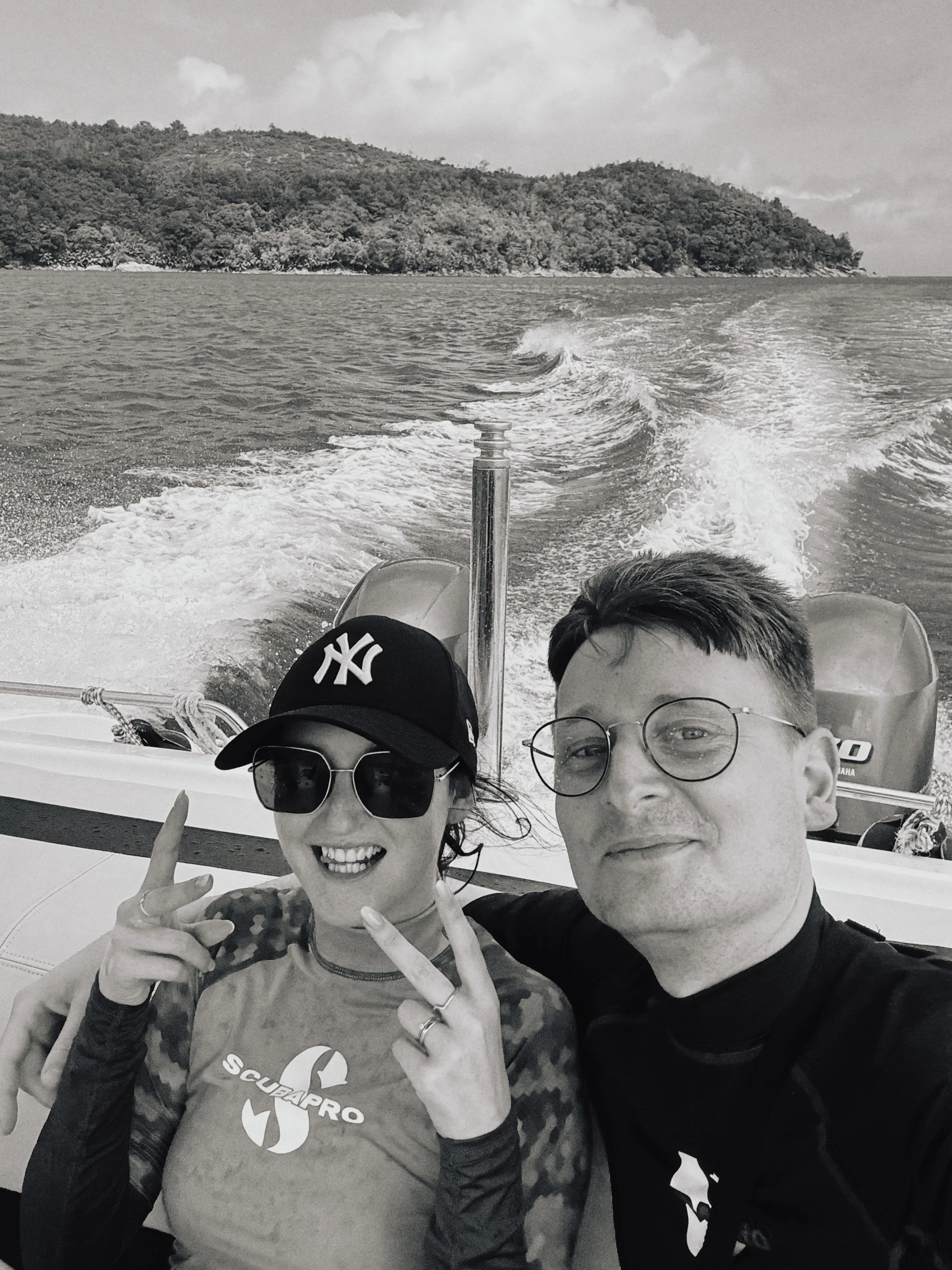 Zwei Menschen posieren auf einem Boot, hinter ihnen ist das Meer und eine Insel mit Bäumen. Es ist schwarz-weiß.