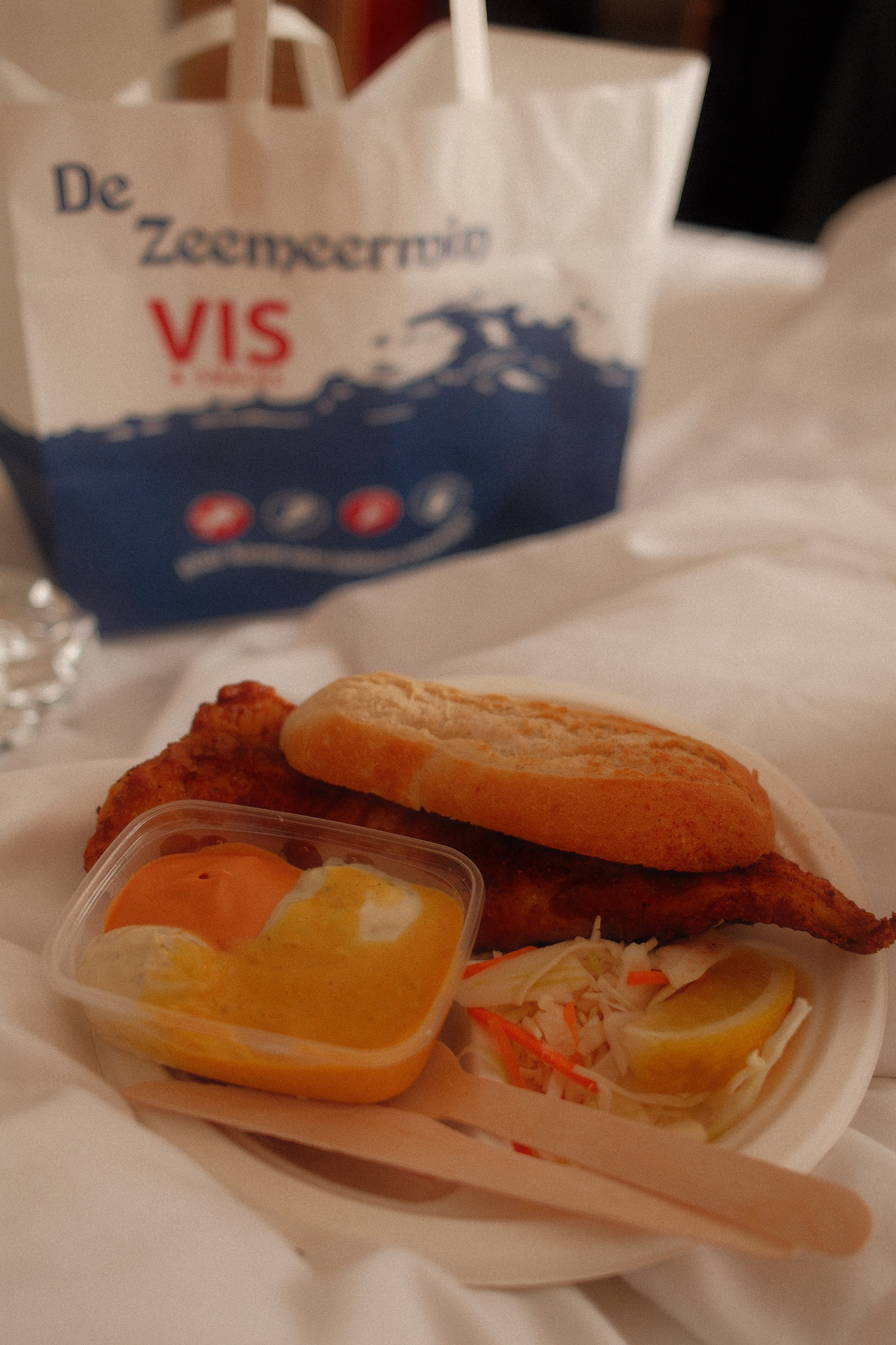 backfischbrötchen mit sauce, krautsalat und zitrone auf einem hotelbett, im hintergrund eine papiertüte von de zeemeermin vis.