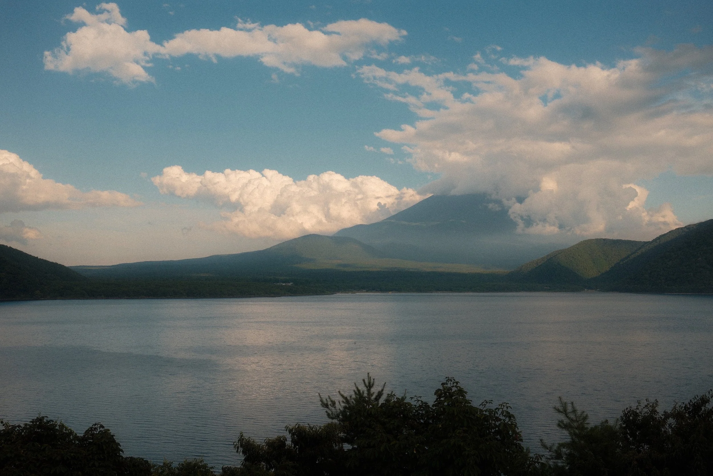 Blick auf einen See mit Bergen im Hintergrund, darunter ein Vulkan, teilweise von Wolken bedeckt, bei Sonnenuntergang oder -aufgang.