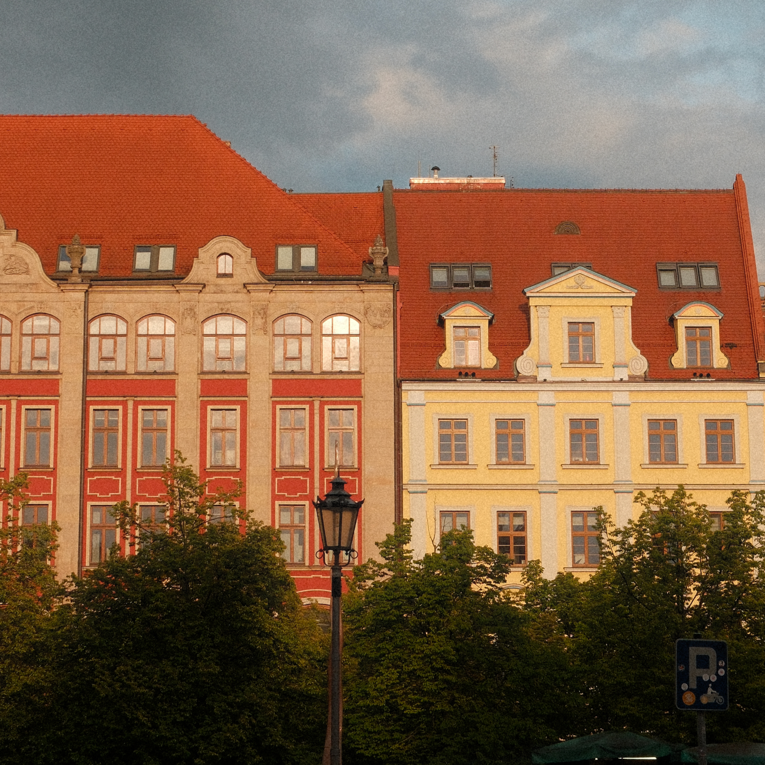 nahaufnahme zweier sanierter altbaufassaden in rot und gelb mit steilen roten ziegeldächern; im vordergrund bäume und eine straßenlaterne, darüber bewölkter himmel im warmen abendlicht.